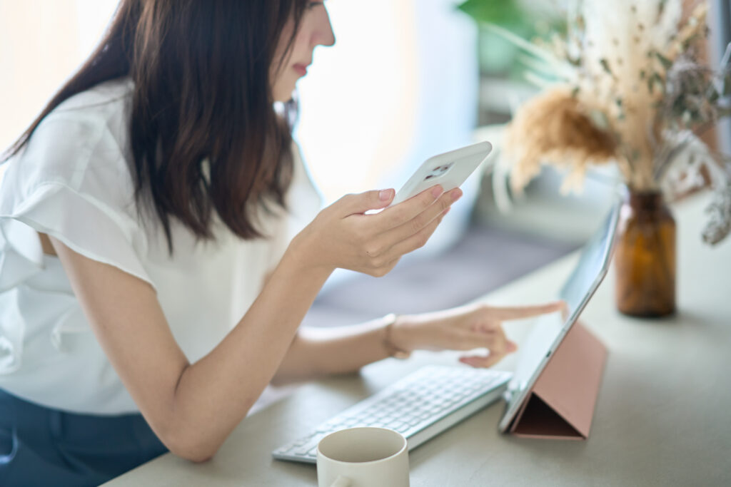 PC画面を前に、スマートフォンを操作する女性
