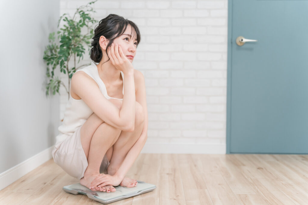 体重計の上で膝を抱えてしゃがみ込み、浮かない表情で考え事をする若い女性